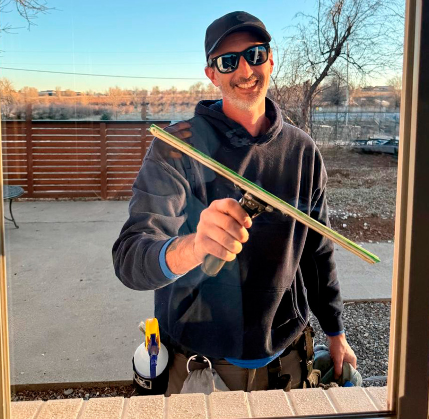 Smiling window cleaning technician using a squeegee on a residential home in Lafayette, Colorado, as sunlight reflects off the glass.